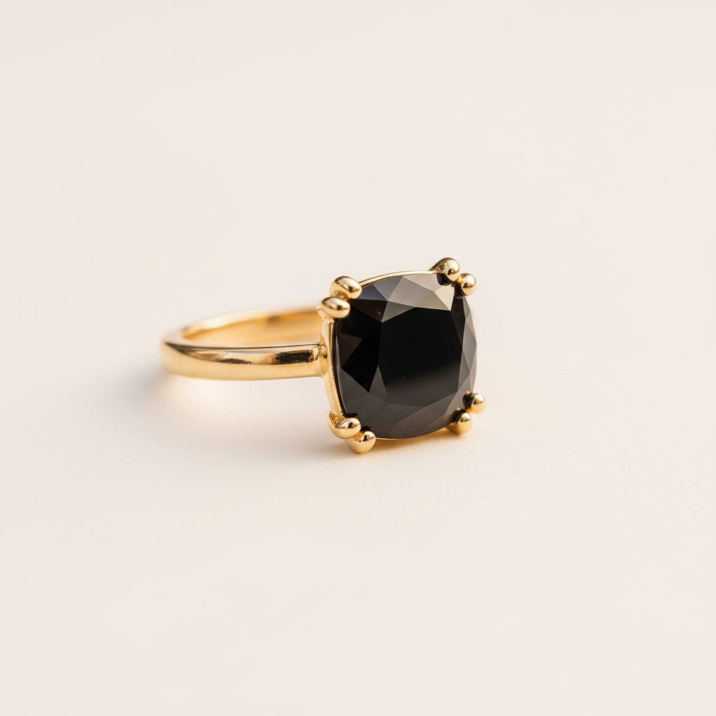 A gold ring with a cushion-cut black gemstone in a six-prong setting, resting on a textured stone surface with a blurred green garden background.