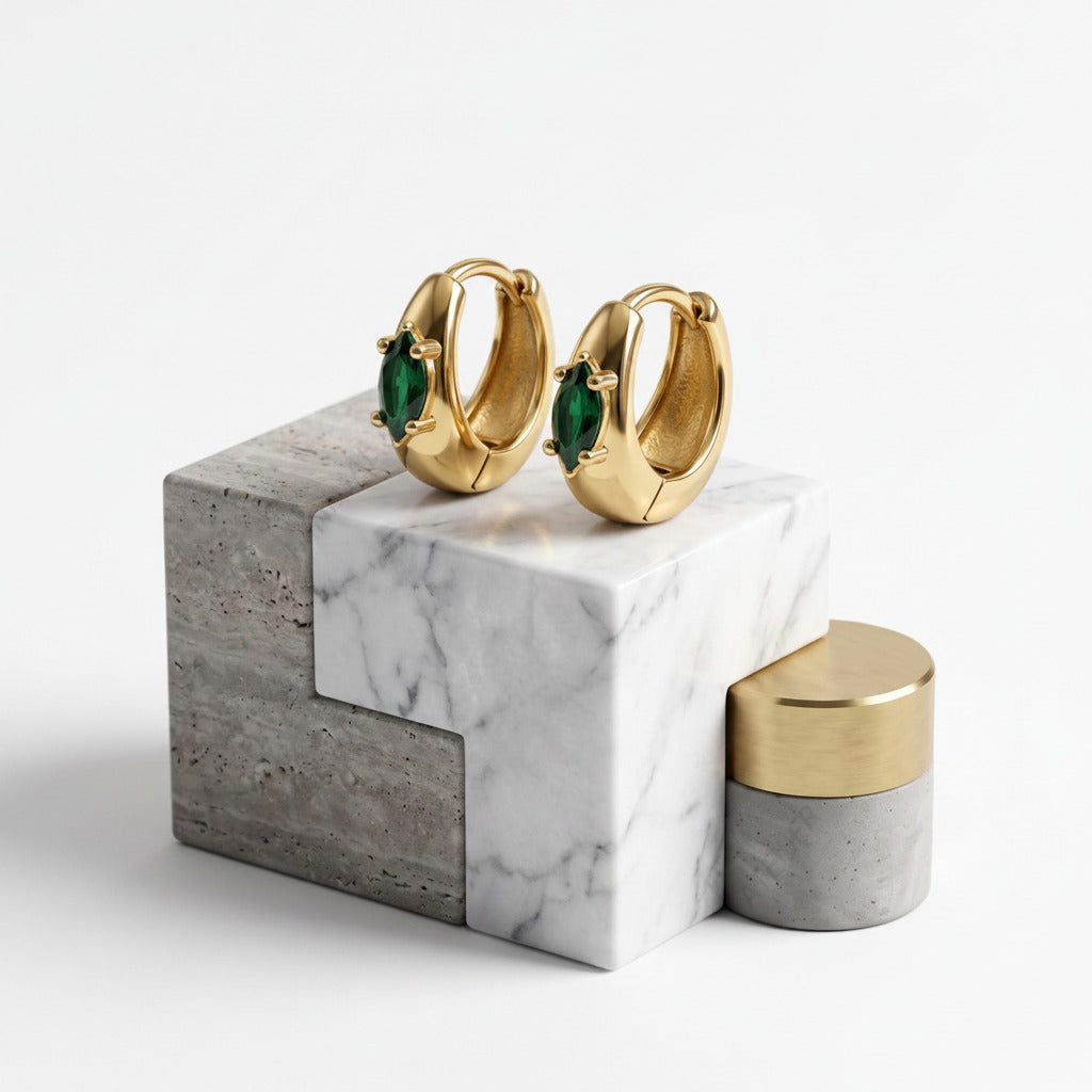 A pair of gold huggie hoop earrings with green marquise-cut gemstones, displayed on a modern gray and white marble block next to a matching gold and gray cylindrical container. The image has a clean white background with diagonal 'MINIMAL ATELIER' watermarks.