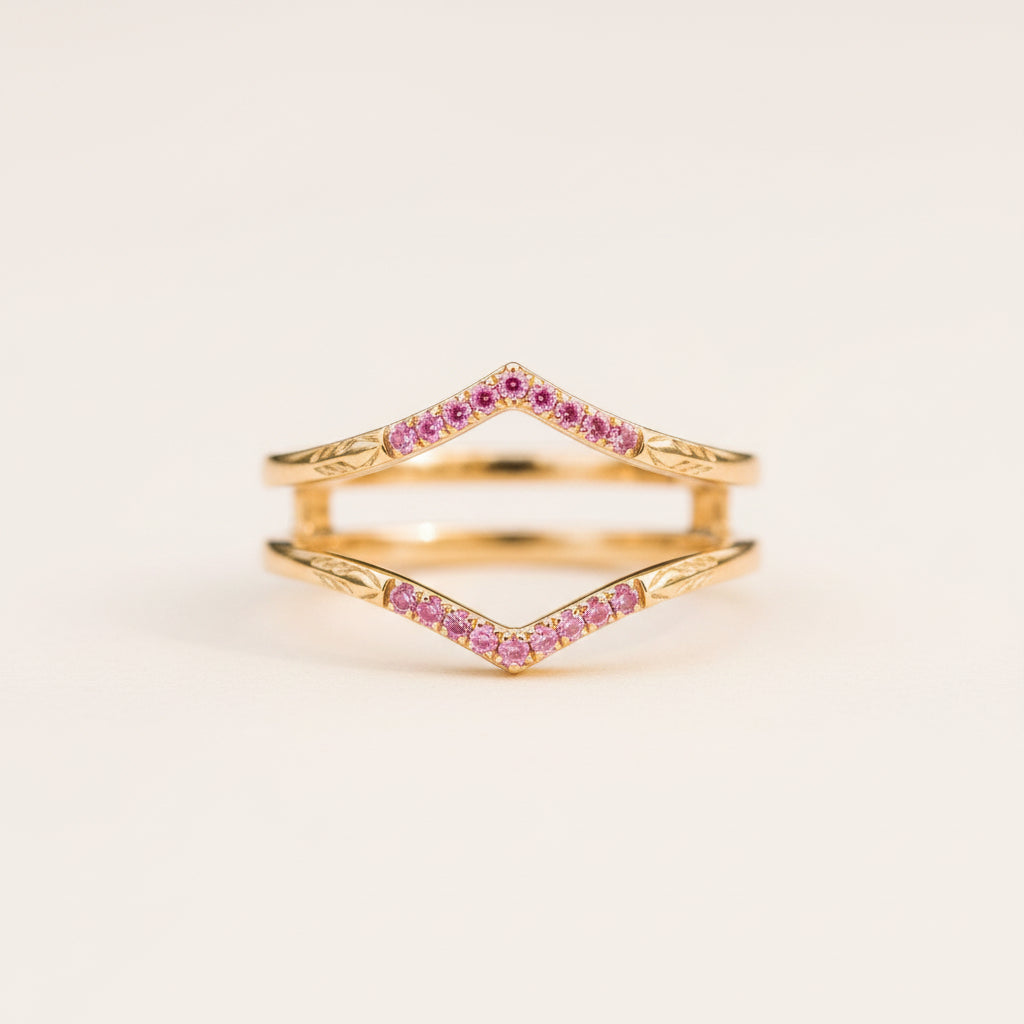 A pair of gold rings with a chevron design set with small pink gemstones, resting on a textured stone surface with a blurred natural background.
