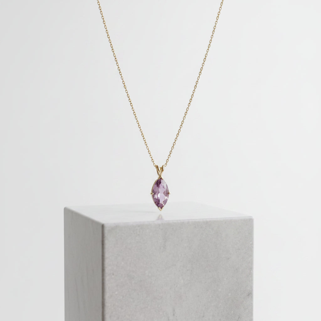 A delicate gold chain necklace with a small teardrop-shaped purple gemstone pendant, displayed on a light gray stone pedestal in a softly lit interior setting.