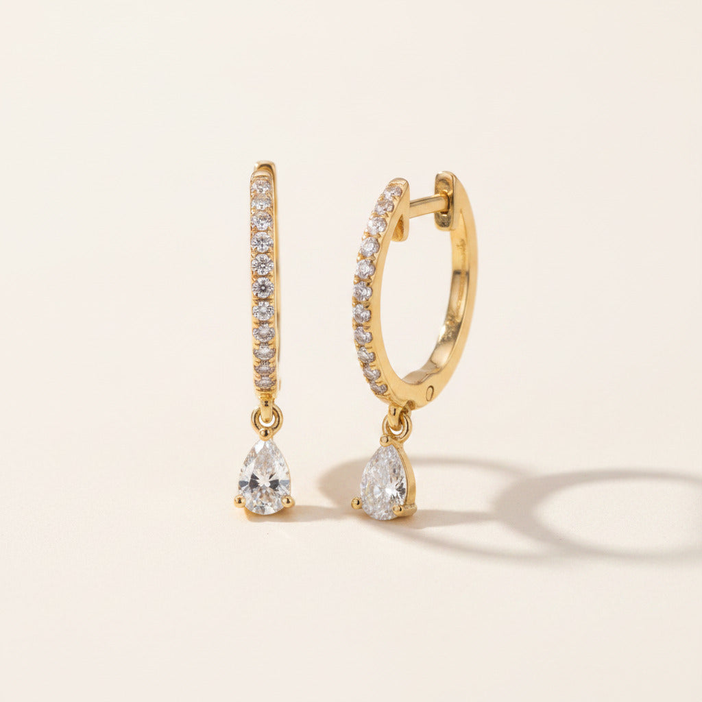 A pair of gold huggie hoop earrings with pavé-set clear stones and pear-cut crystal drop charms, displayed on a textured navy blue velvet surface.