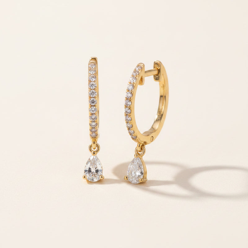 A pair of gold huggie hoop earrings with pavé-set clear stones and pear-cut crystal drop charms, displayed on a textured navy blue velvet surface.