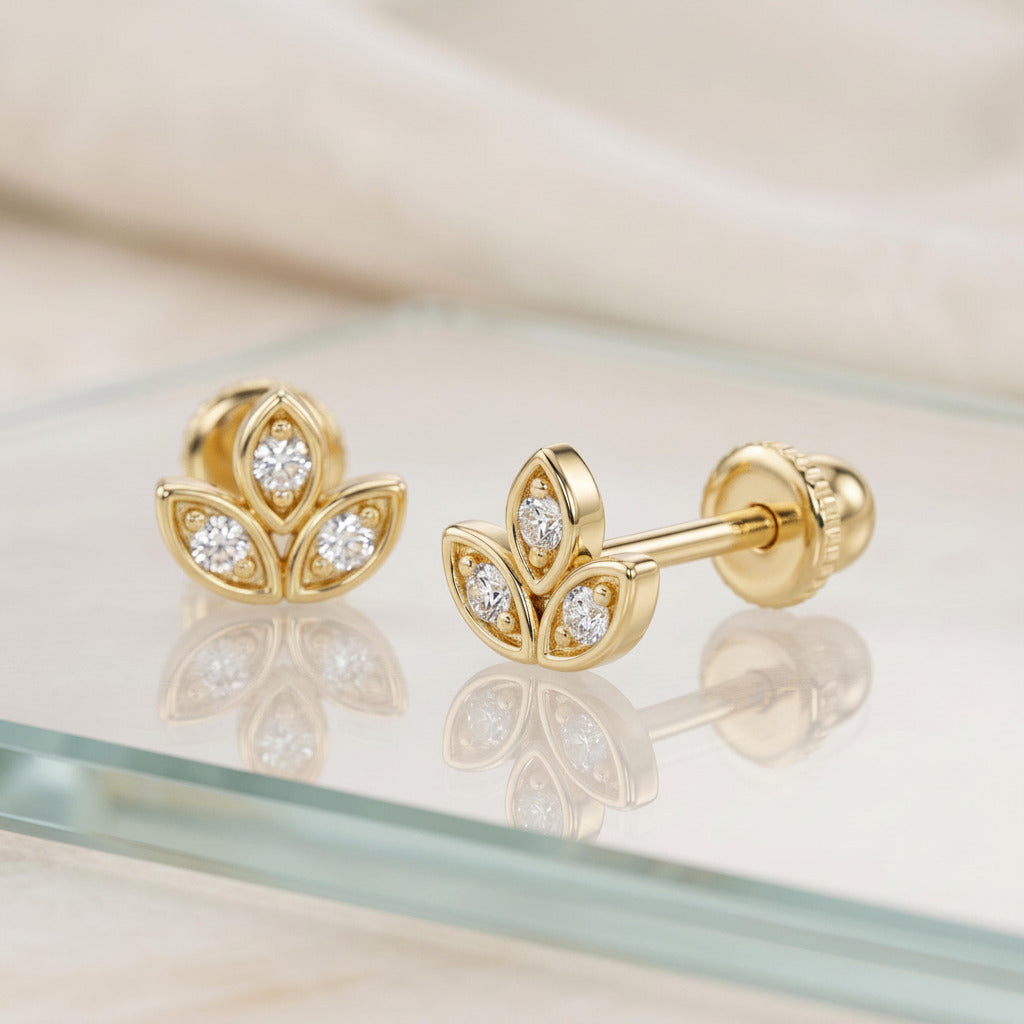 A pair of gold flower-shaped stud earrings with clear gemstones, displayed on a glass surface with a soft beige fabric background.