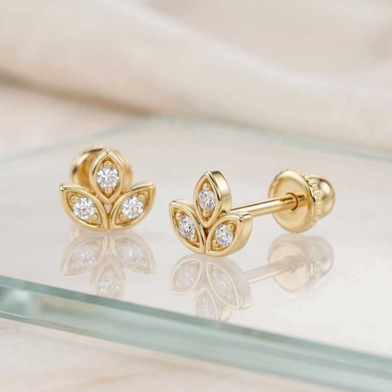 A pair of gold flower-shaped stud earrings with clear gemstones, displayed on a glass surface with a soft beige fabric background.