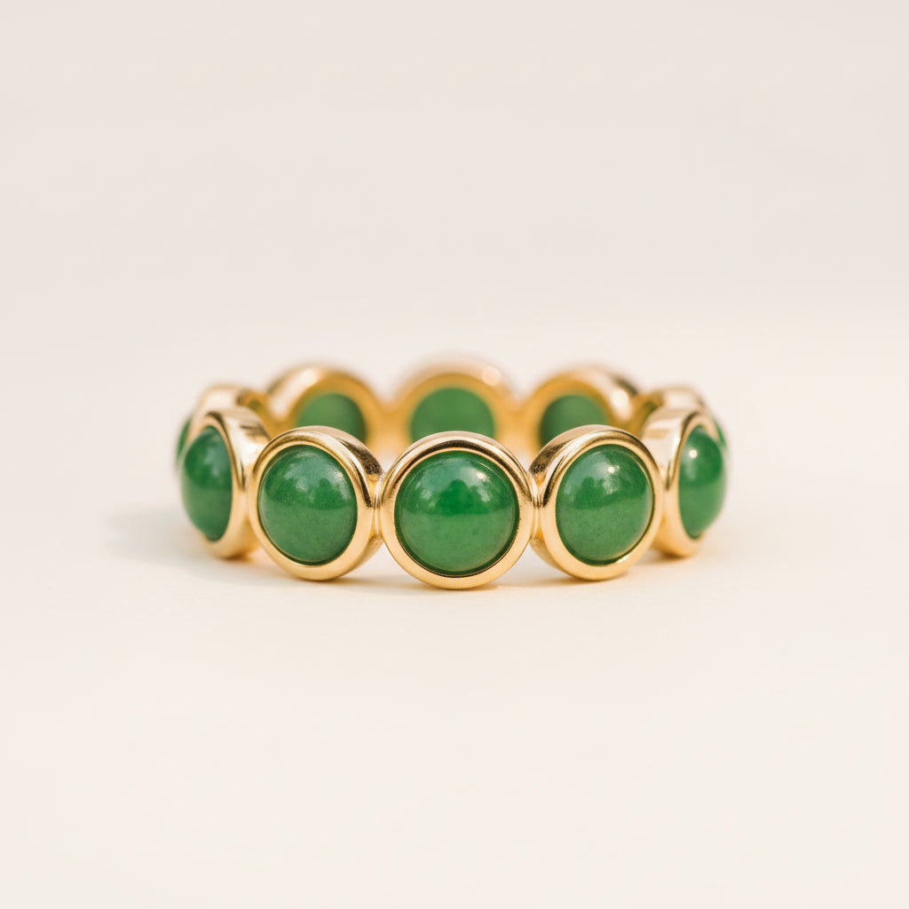 A gold band ring featuring multiple round green jadeite gemstone bezels, resting on a textured stone surface with a blurred natural background.