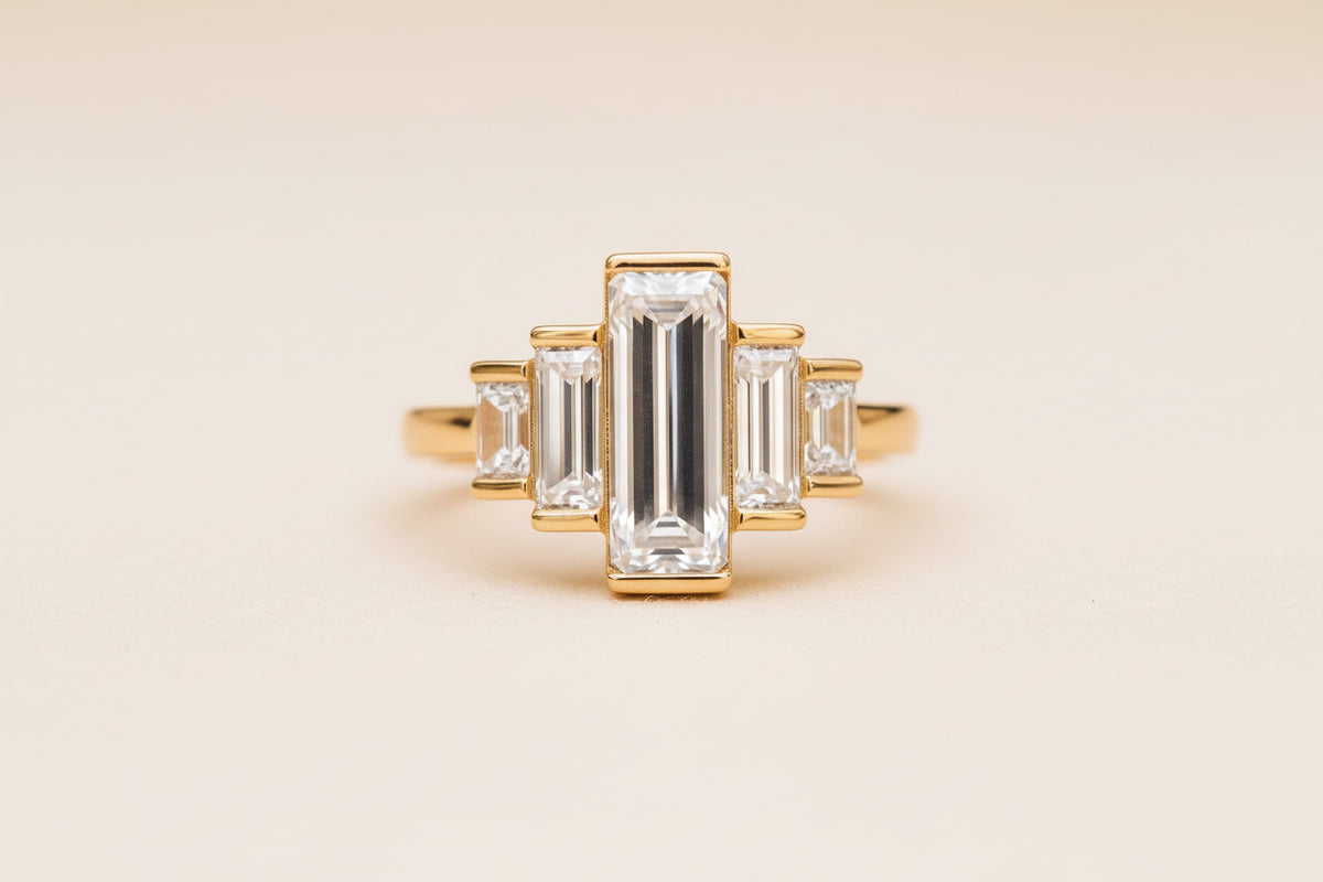 A gold ring featuring a central rectangular baguette-cut clear gemstone flanked by smaller matching baguette and round-cut stones, photographed resting on a textured stone surface with a softly blurred natural background.