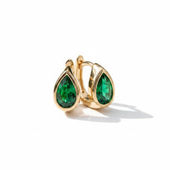 A pair of gold hoop earrings with teardrop-shaped green gemstones, resting on a textured light gray stone surface.