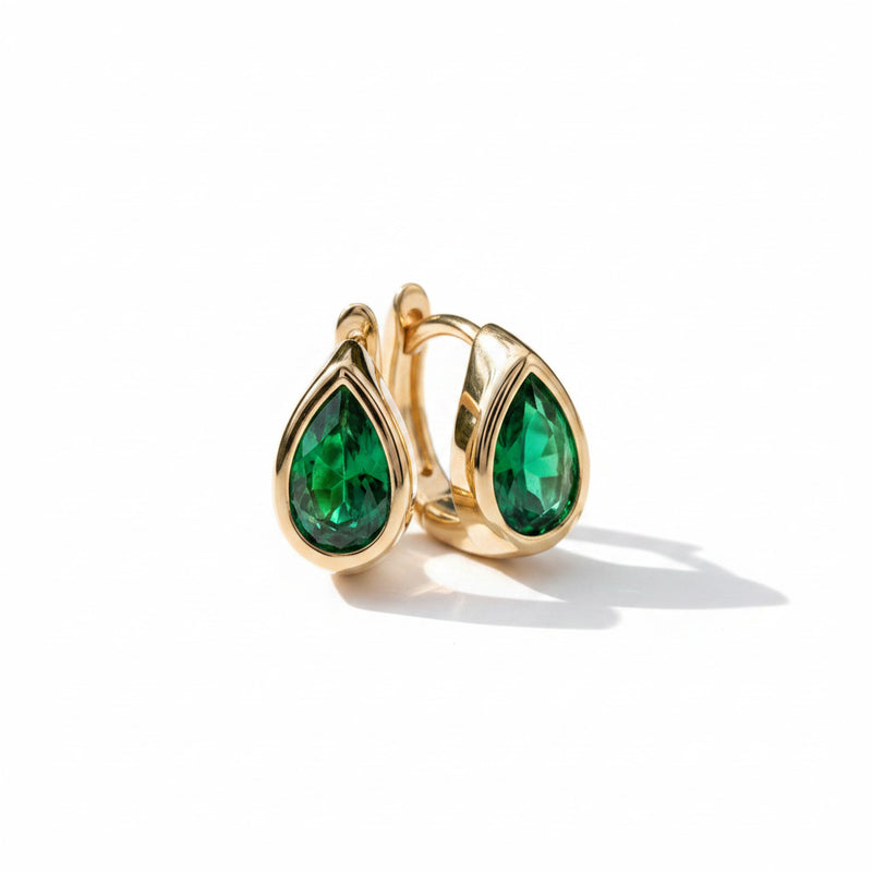 A pair of gold hoop earrings with teardrop-shaped green gemstones, resting on a textured light gray stone surface.