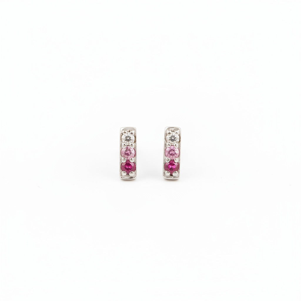 A pair of silver-toned rectangular stud earrings with pink gemstones, resting on a textured piece of weathered wood.
