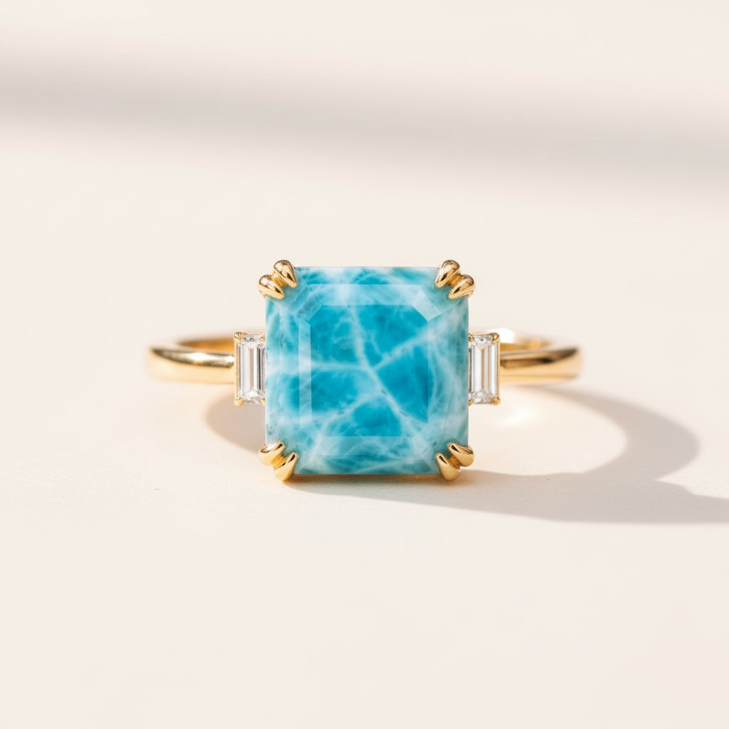 A gold ring featuring a square-shaped light blue larimar gemstone with white veining, accented by baguette side stones, resting on a textured stone surface with a blurred natural background.