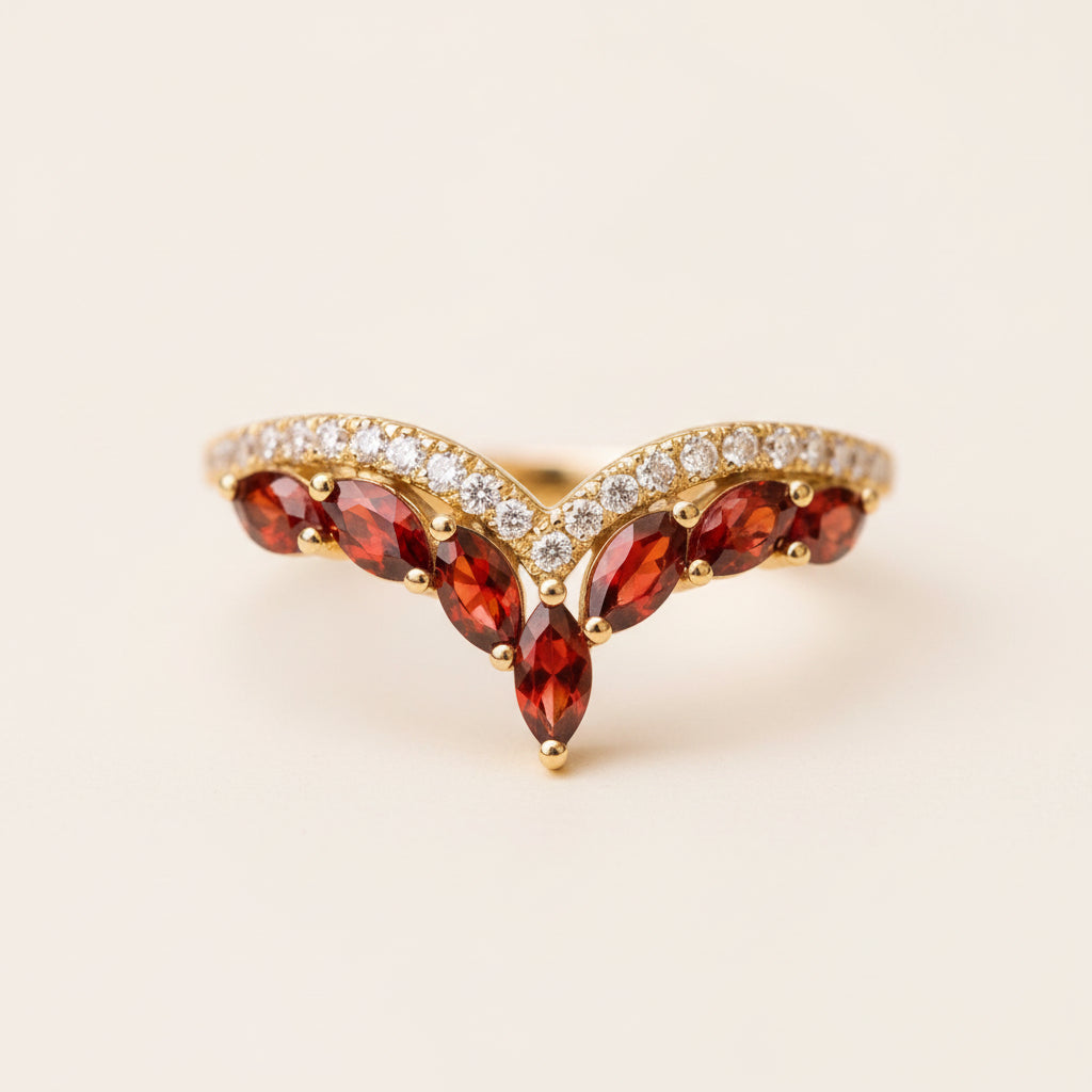 A gold ring with marquise-cut red gemstones and a row of small clear stones, photographed close up on a textured stone surface with a blurred natural background.