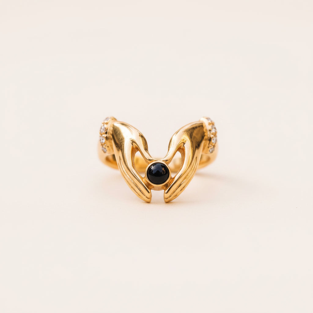 A gold ring shaped like two embracing hands, featuring a central round black gemstone and small clear stones along the sides, resting on a textured stone surface with a blurred natural background.