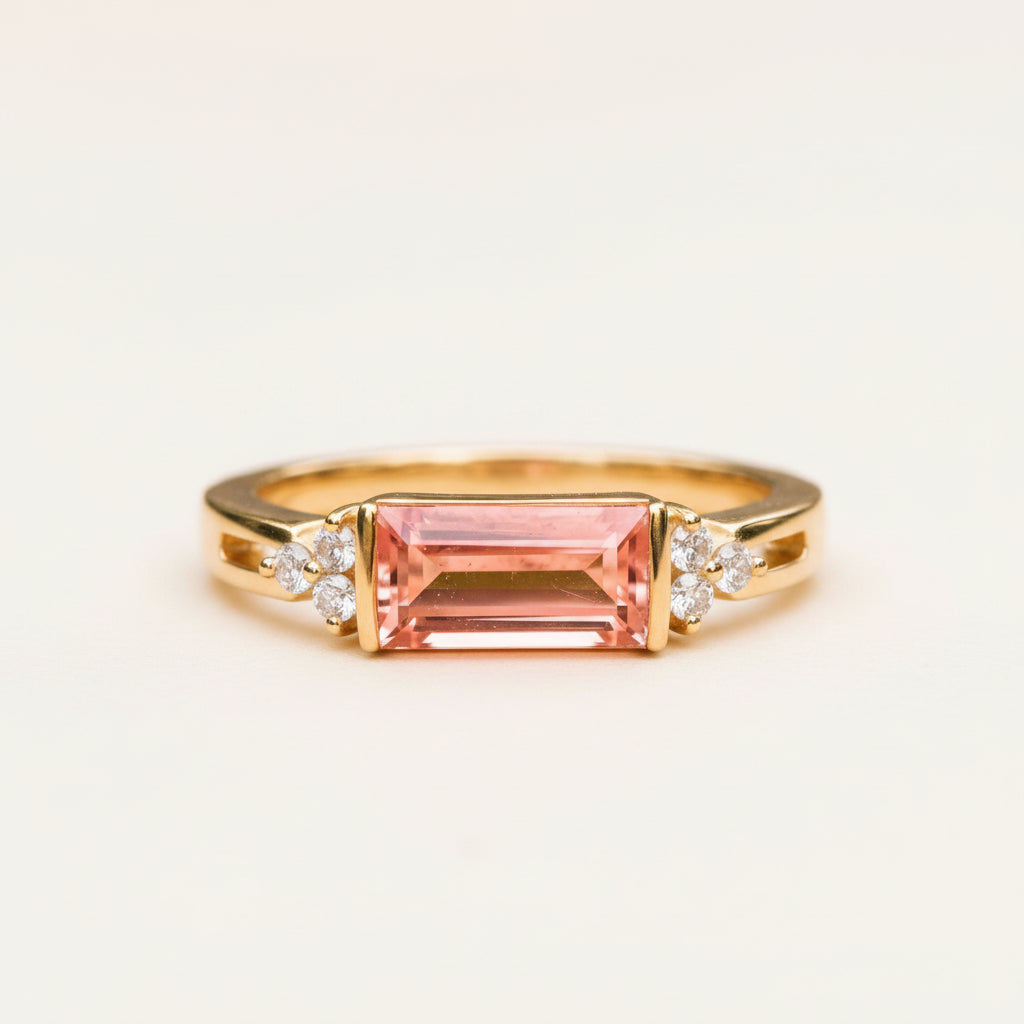 A yellow gold ring featuring a rectangular emerald-cut pink gemstone, accented by small diamond clusters on either side, resting on a textured stone surface with a blurred natural background.