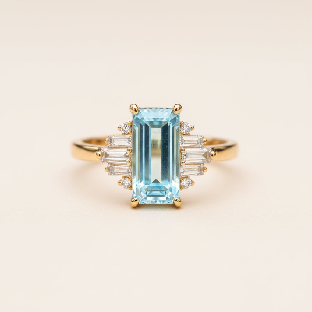 A gold ring featuring a large emerald-cut light blue gemstone, flanked by baguette and round clear stones, resting on a textured wooden surface with a blurred natural background.