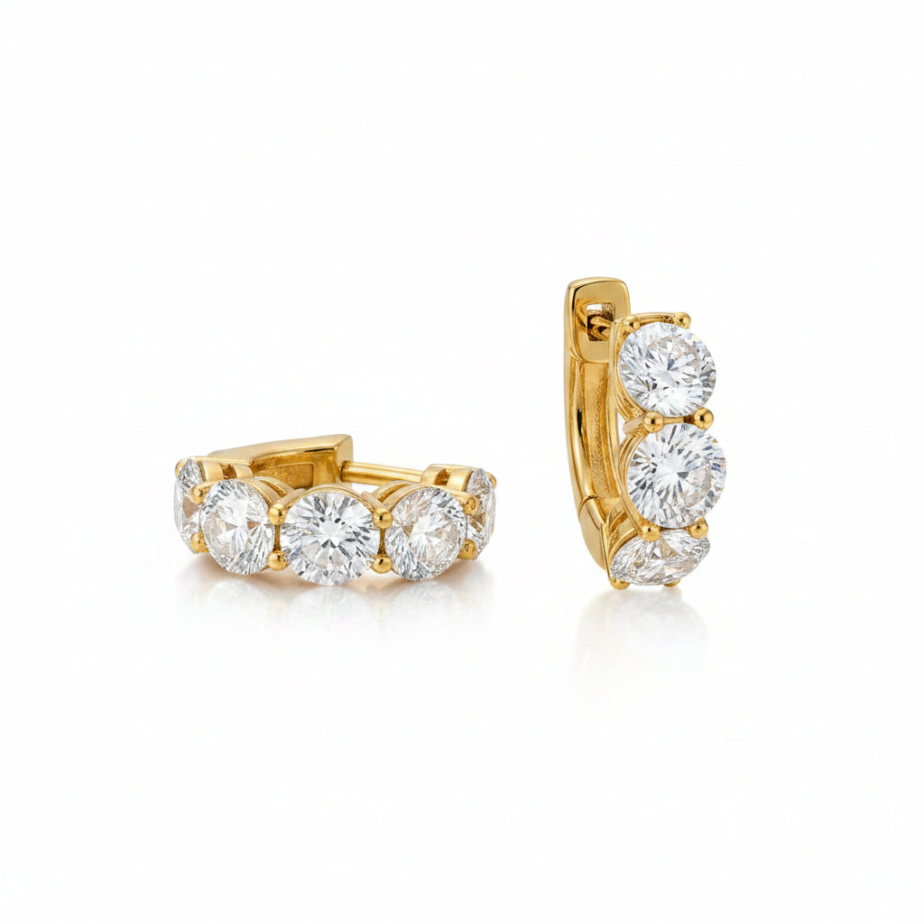 A pair of gold huggie hoop earrings with clear gemstones, displayed on a light beige marble slab against a warm brown background.