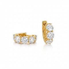 A pair of gold huggie hoop earrings with clear gemstones, displayed on a light beige marble slab against a warm brown background.