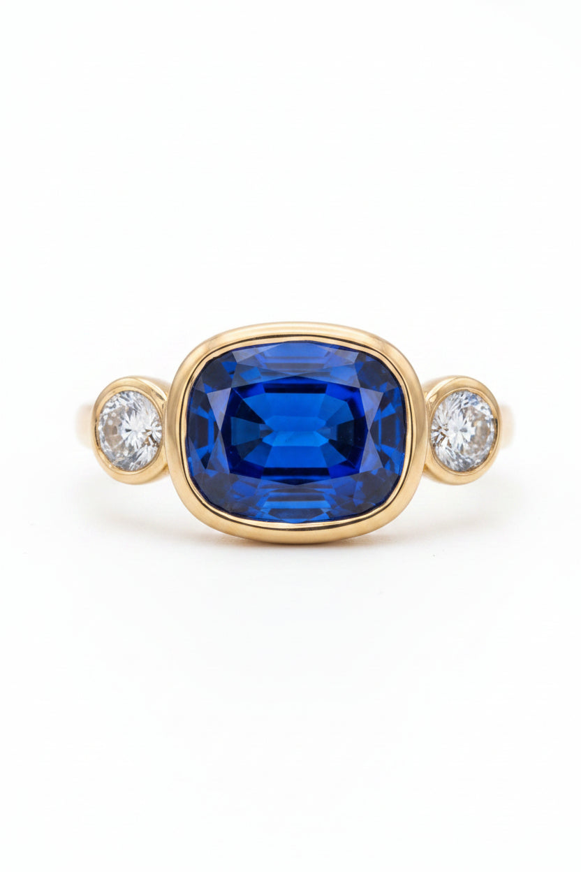 A yellow gold ring featuring a large cushion-cut deep blue sapphire center stone, flanked by two round brilliant-cut diamonds in gold bezels, set against a dark black velvet background.