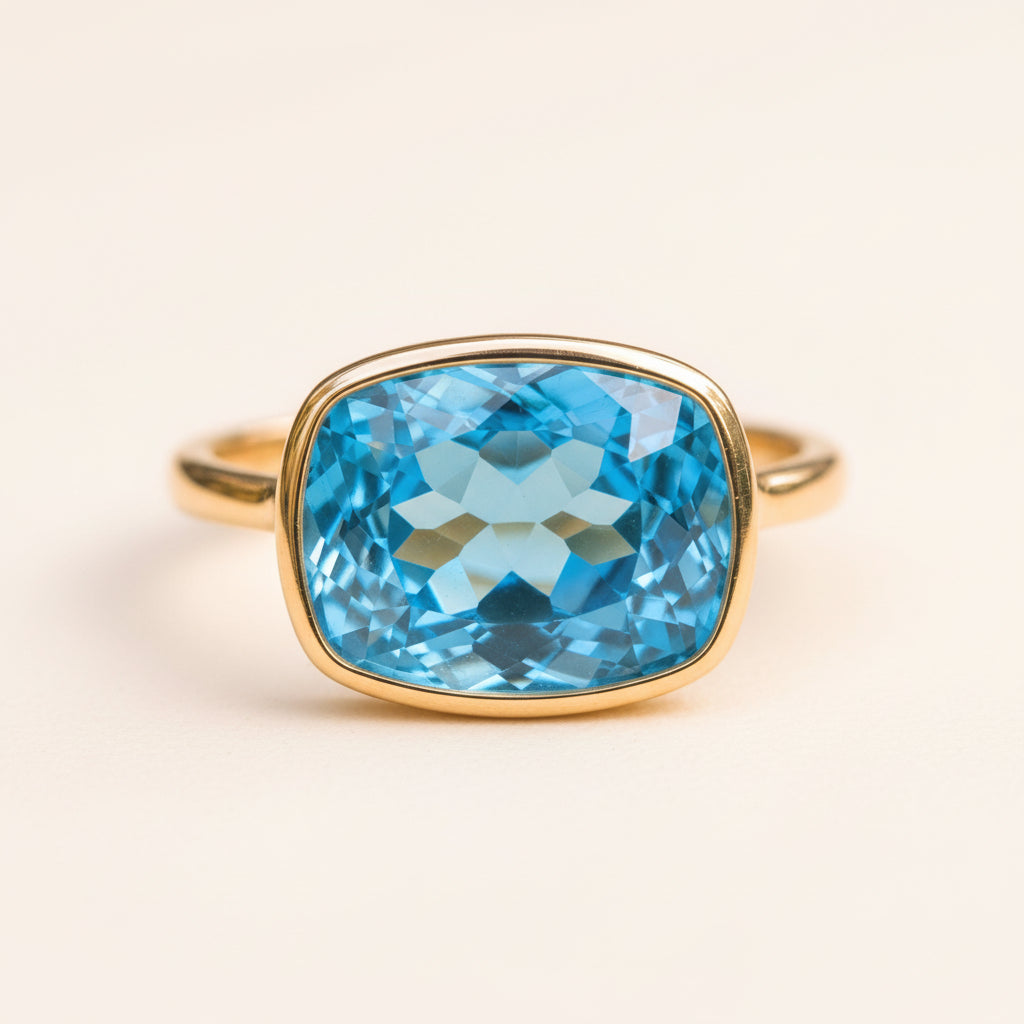 A cushion-cut blue topaz gemstone ring in a gold bezel setting, resting on a textured stone surface with a soft-focus natural background.