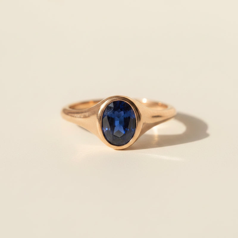 A gold signet ring with an oval-cut deep blue sapphire gemstone, resting on a textured beige linen fabric under warm, directional sunlight.