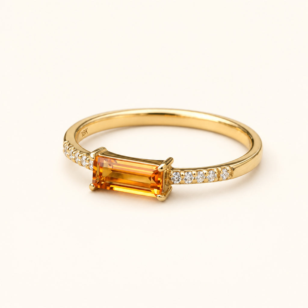 A gold ring with a rectangular amber-orange center stone and small clear accent stones on the shoulders, resting on a textured stone surface with a blurred natural background.