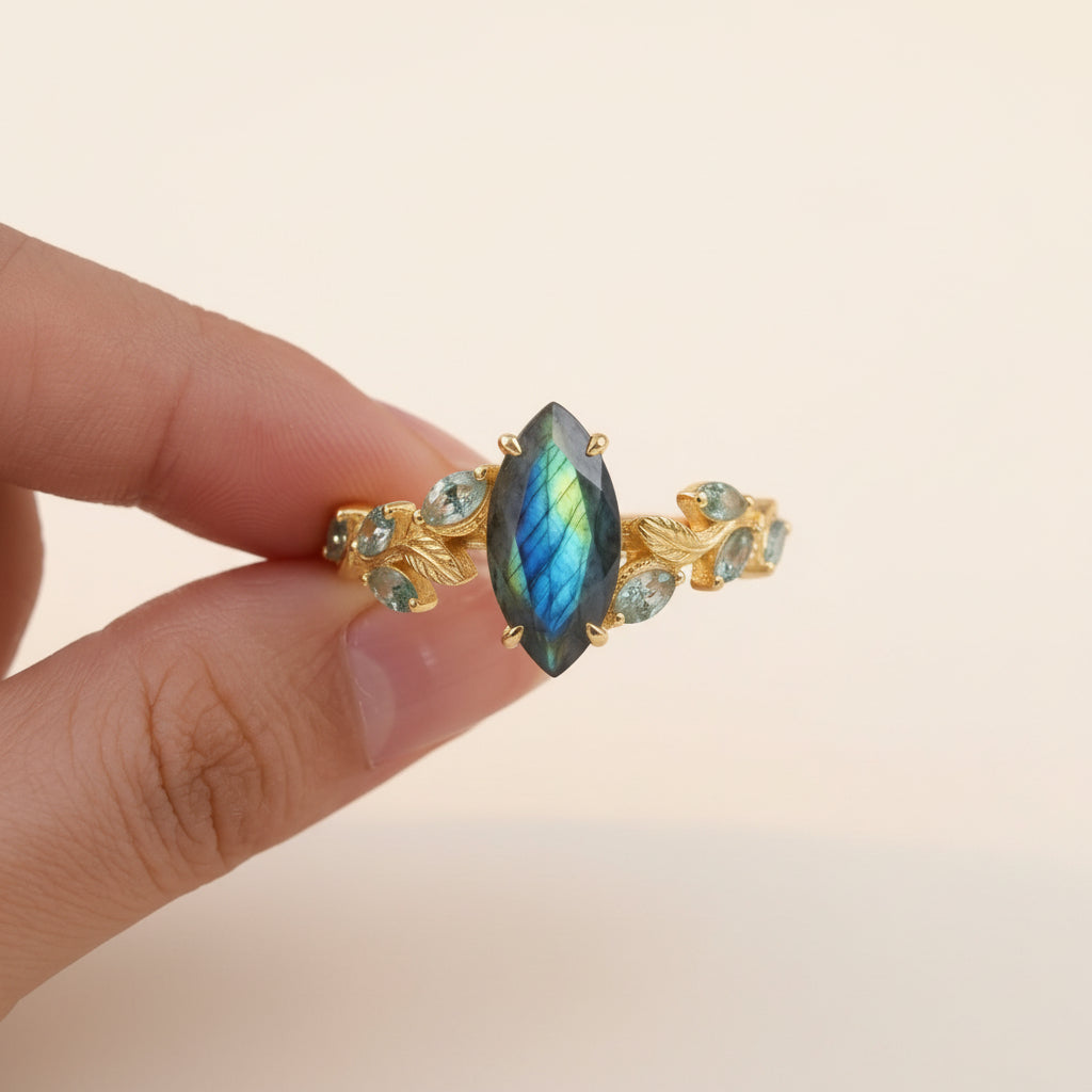 A gold ring with a marquise-shaped iridescent blue-green labradorite stone and small green stones on either side, held between fingers against a soft cream lace background with dried flowers.