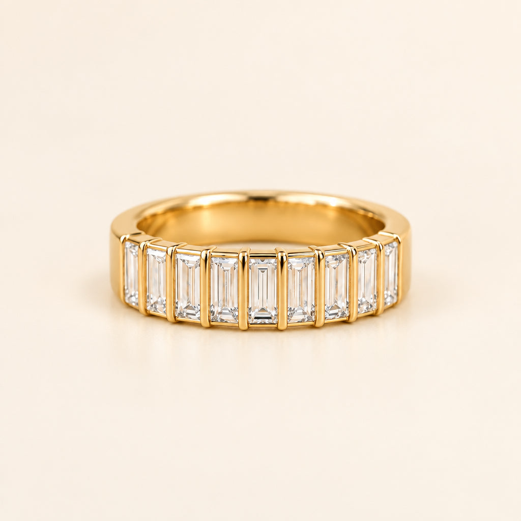 A gold band ring featuring a row of baguette-cut clear gemstones, displayed on draped cream-colored satin fabric.