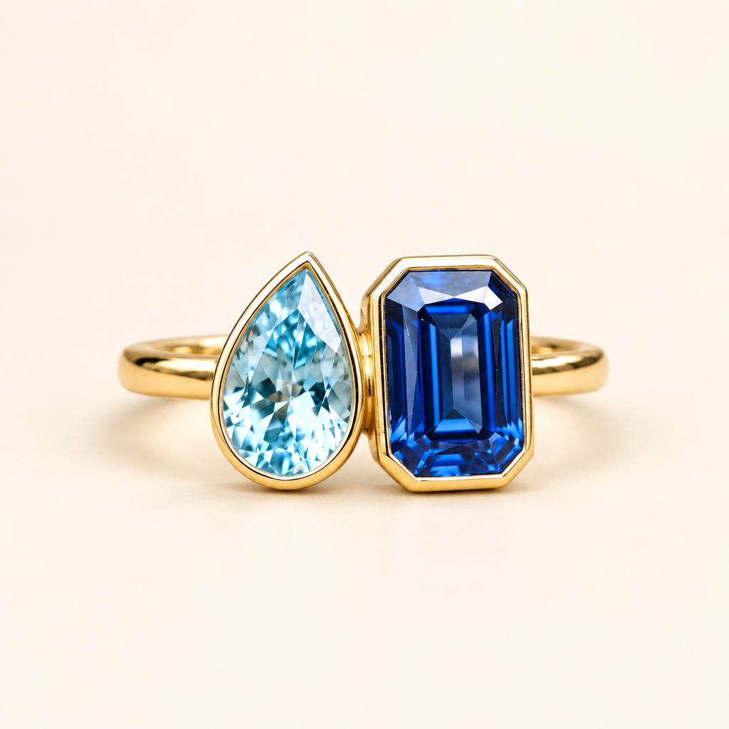 Two gold rings displayed on a textured wood surface with a blurred natural background. One ring features a teardrop-shaped light blue gemstone, and the other features a rectangular deep blue gemstone, both set in yellow gold.