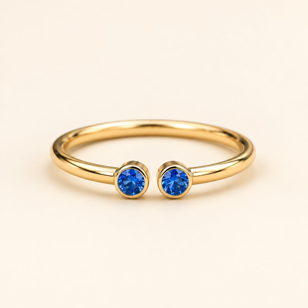 A gold open-bend ring with two round blue gemstones, resting on a textured stone surface with a blurred natural background. Watermarked with 'GoldGemsGlitter'.