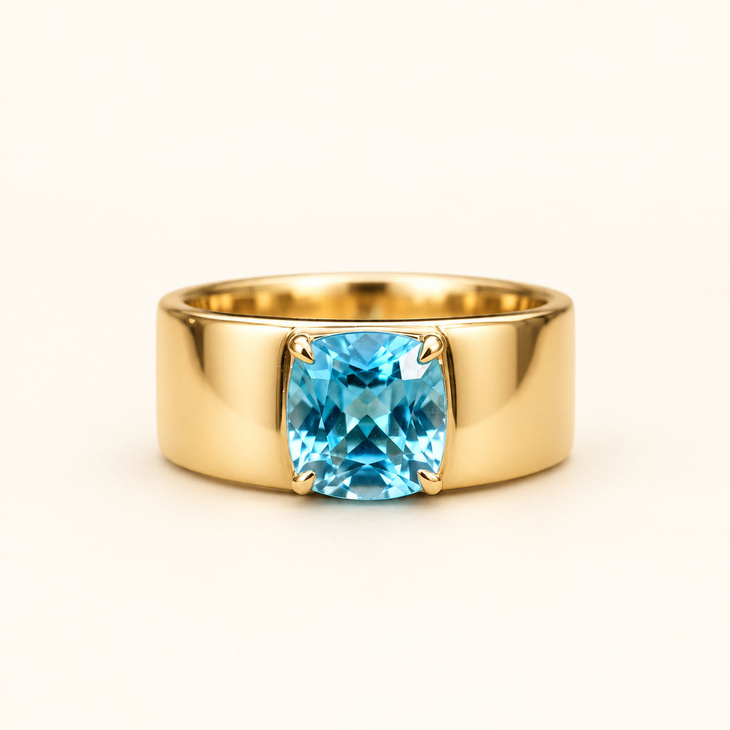 A gold ring with a cushion-cut light blue gemstone, resting on a textured stone surface with a blurred natural background.