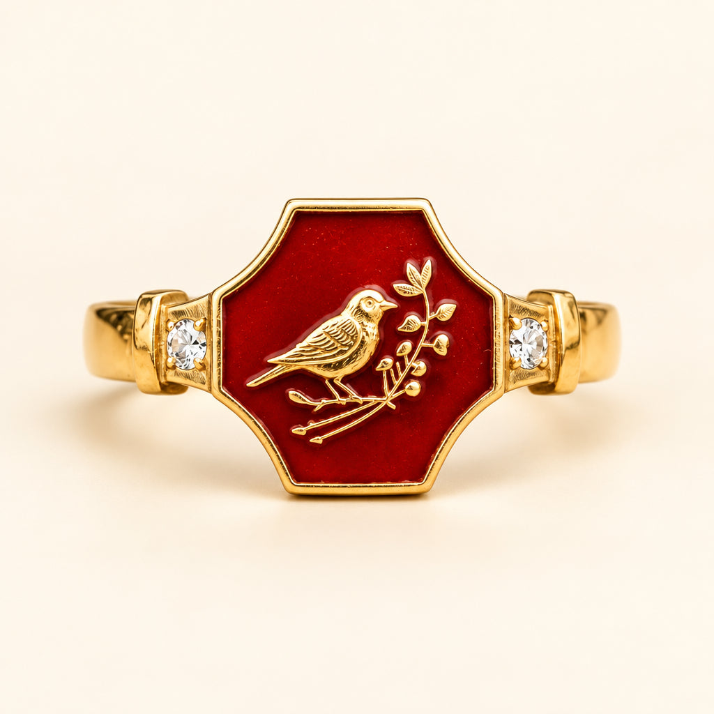 A gold signet ring with a hexagonal red enamel face featuring an engraved bird and branch motif, displayed on a textured stone surface with a blurred natural background.