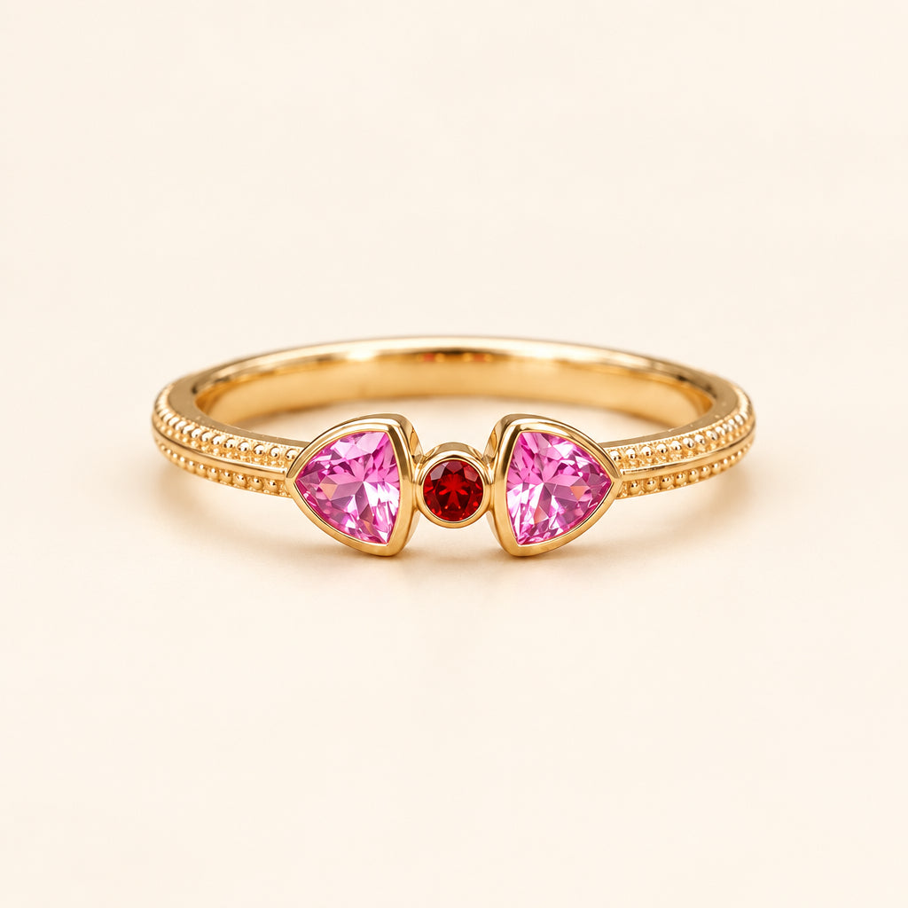 A gold ring with two pink triangular gemstones and a small red center stone, resting on a textured stone surface.