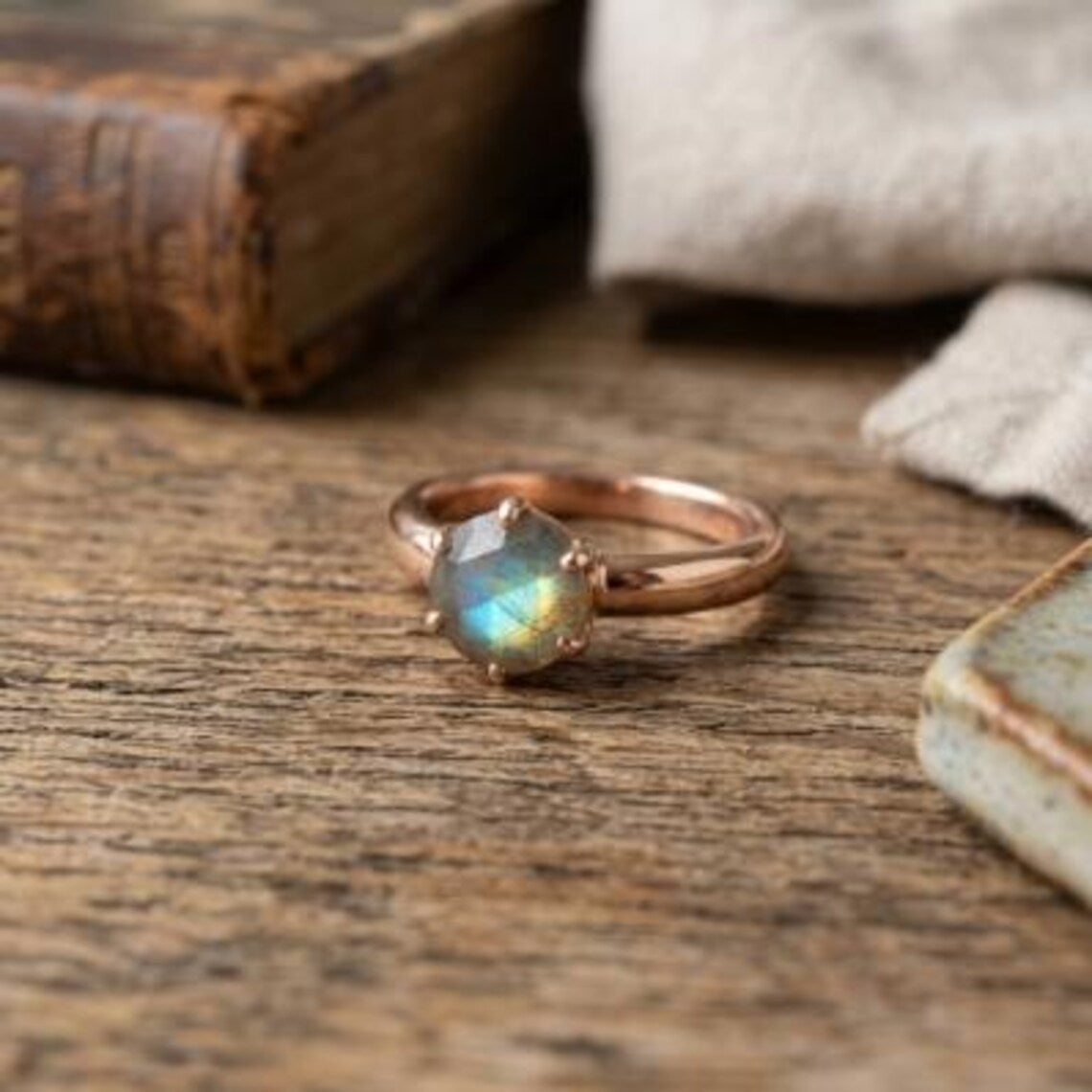 A rose gold ring with a faceted iridescent labradorite gemstone, resting on a rustic wooden surface with a blurred vintage book and fabric in the background.