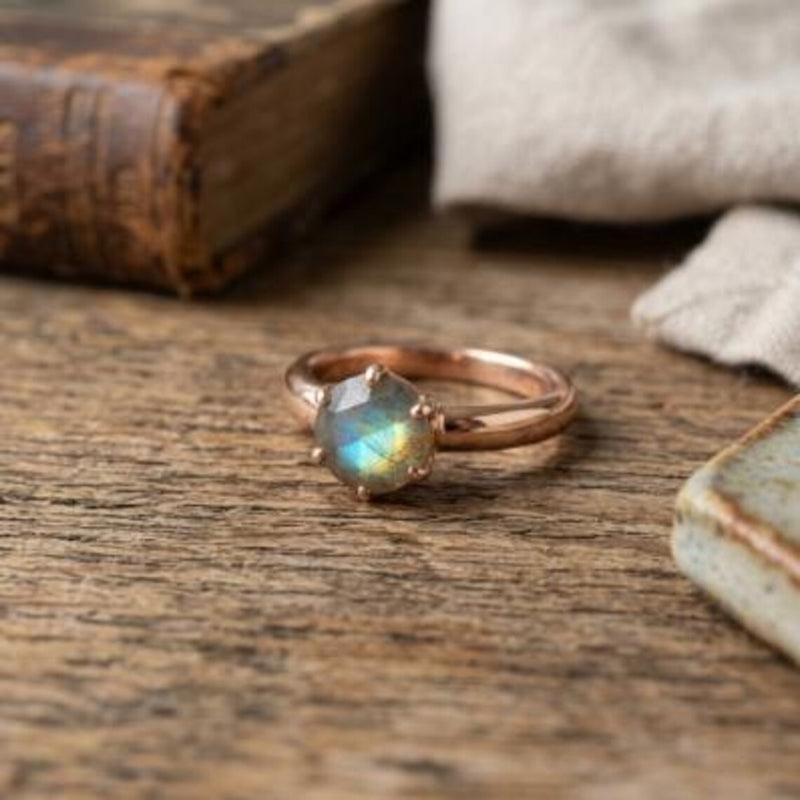 A rose gold ring with a faceted iridescent labradorite gemstone, resting on a rustic wooden surface with a blurred vintage book and fabric in the background.