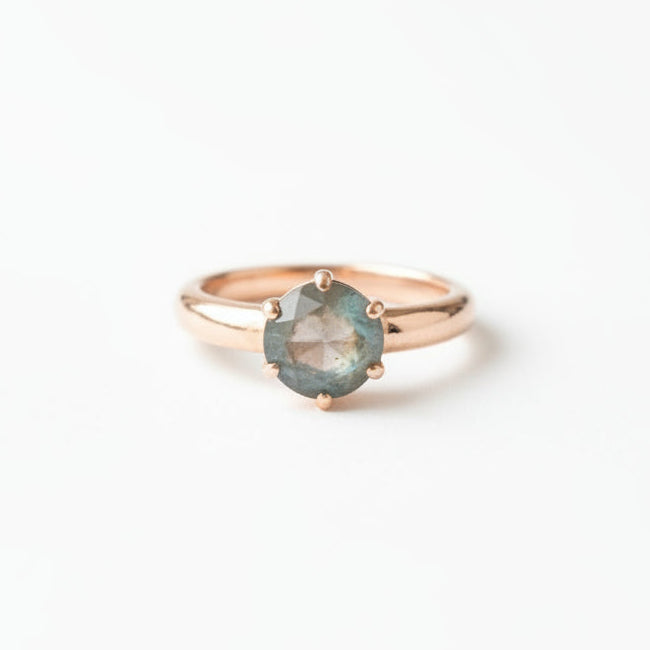 A delicate gold ring with a round iridescent blue-green opal gemstone, resting on a textured brown leather surface.