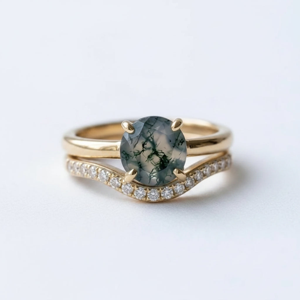 A gold ring set featuring a round greenish-gray stone with dark veining, accented by a wavy pavé band with small clear stones, resting on a soft white fabric surface.