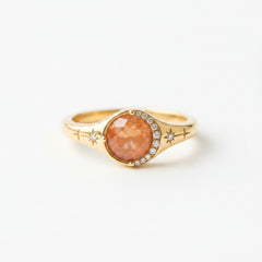 A gold signet ring with a round, faceted orange-brown gemstone and small clear accent stones, resting on a rustic wooden surface.