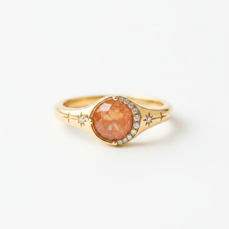 A gold signet ring with a round, faceted orange-brown gemstone and small clear accent stones, resting on a rustic wooden surface.